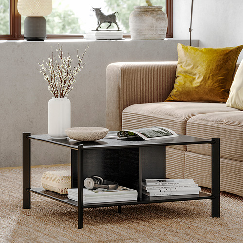 Sala de estar moderna con sofá beige, almohadas mostaza, mesa de centro negra. La mesa tiene estantes de bambú con libros, jarrones, artículos decorativos.