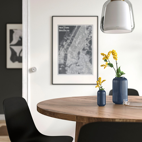 Comedor moderno con mesa de madera, sillas negras, dos jarrones azules con flores amarillas, póster en blanco y negro, luz colgante.