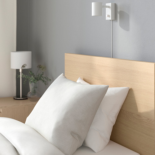 Una estructura de cama de madera con un cabecero alto y almohadas contra una pared. Una lámpara blanca junto a un jarrón con vegetación se coloca en la mesita de noche de madera.