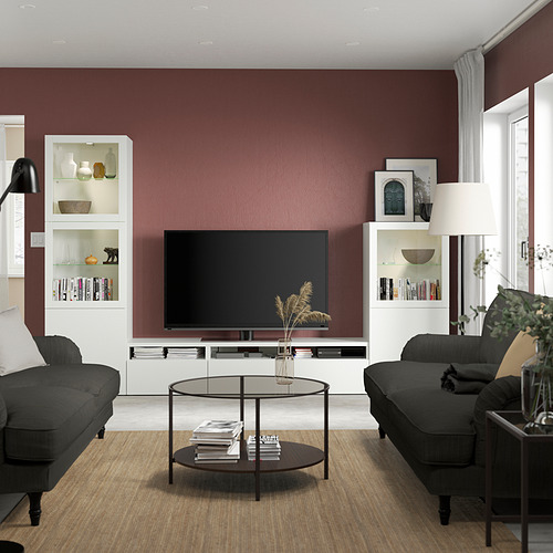 Sala de estar moderna con paredes de color rojo oscuro, mueble de TV BESTÅ blanco, TV de pantalla plana, sofá gris, mesa de centro de cristal, lámpara de pie y plantas.