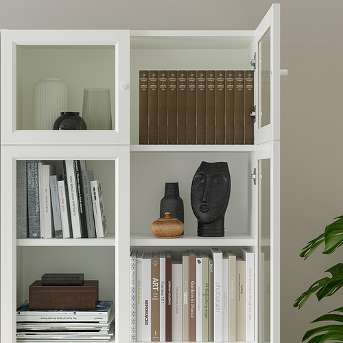 Estantería blanca con estantes, que exhibe libros y artículos decorativos como jarrones y esculturas. Puertas de vidrio y panel disponibles.