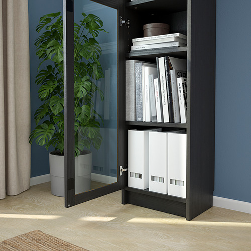 Mueble moderno de madera oscura con puerta de cristal, que alberga libros y carpetas. A su lado hay una planta alta en maceta.