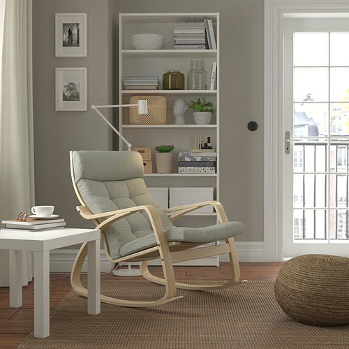Mecedora POÄNG en un entorno tranquilo. Gris claro, respaldo alto, con mesa auxiliar y estantería. La decoración neutra incluye otomana y cortinas.
