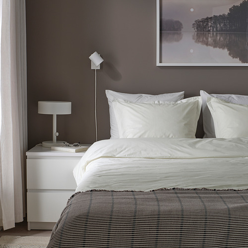 Cama hecha con edredón HAMPFLOCKEL blanco, tejido de algodón nítido. Pared gris, mesita de noche blanca con lámpara.