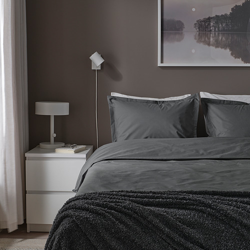 Dormitorio minimalista con ropa de cama HAMPFLOCKEL gris oscuro, que incluye edredón y 2 almohadas, sobre una mesita de noche blanca. Aplique de pared moderno y pintura.