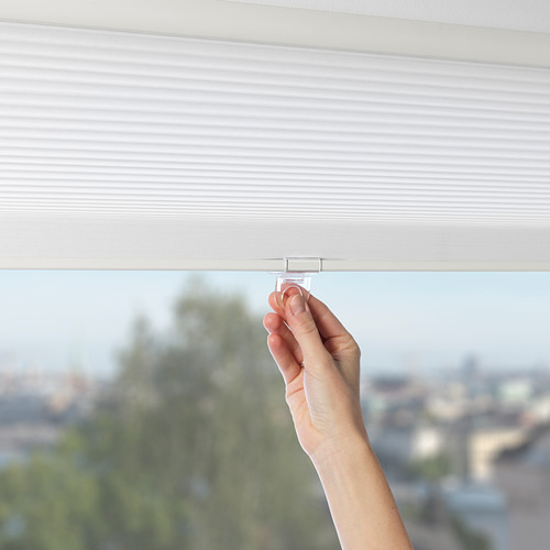 Una mano ajusta una persiana celular blanca en una habitación, controlando la luz solar y la privacidad. El mecanismo de cable oculto garantiza la seguridad.