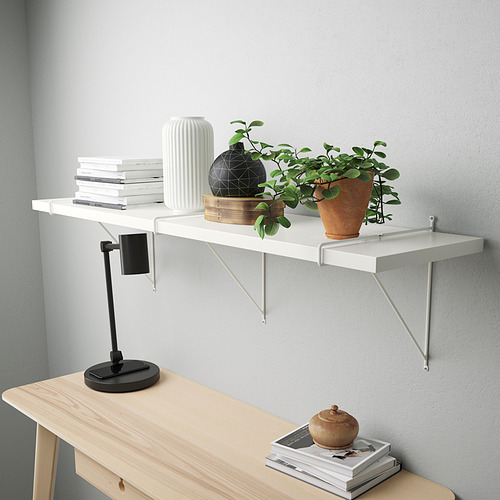 Una estantería de pared con libros y plantas. Tiene un borde biselado y soportes blancos. Debajo hay un escritorio con una lámpara y más libros.