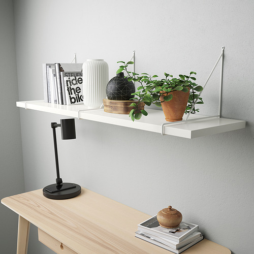 Estante de pared blanco moderno con borde biselado, sosteniendo libros, planta en maceta, jarrón negro. Abajo, una consola de madera clara con lámpara y revistas.
