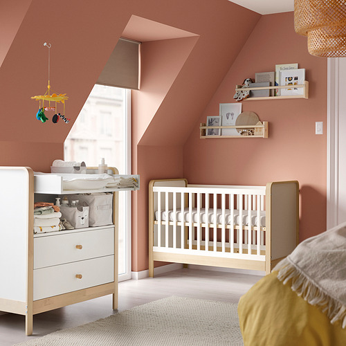 Guardería moderna con paredes rosas, cuna, tocador y cambiador de madera blanca, móvil colgante, estanterías con libros y decoración.