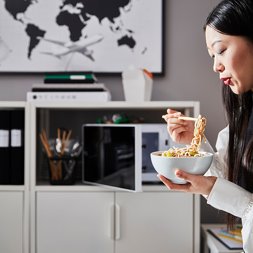 Mujer comiendo fideos en el escritorio de almacenamiento blanco de ROTTEN. El escritorio tiene pies ajustables, almacenamiento con cerradura y construcción de tubo de metal.