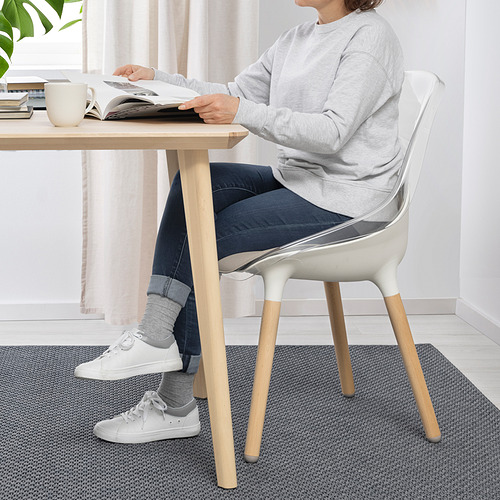 Persona sentada en la mesa leyendo, usando la silla BALTSAR. La silla tiene un asiento transparente, patas de haya robustas y un asiento en forma de cuenco.