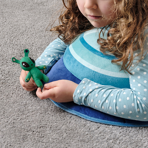Una niña sentada en una alfombra, sosteniendo un pequeño juguete alienígena verde. El juguete tiene un lazo en la parte superior para facilitar su transporte.