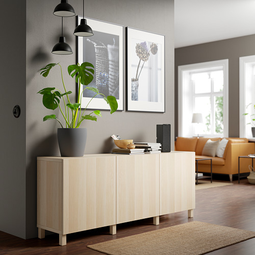 Aparador moderno BESTÅ de madera clara con estantes ajustables, plantas, libros y lámparas. El sofá naranja y la gran ventana crean un espacio acogedor y bien iluminado.