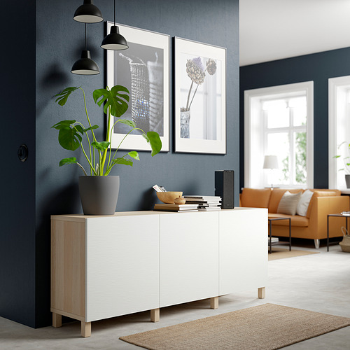 Espacio habitable minimalista con un aparador blanco BESTÅ, con una planta alta, obras de arte enmarcadas y muebles naranjas, frente a una pared gris oscuro.