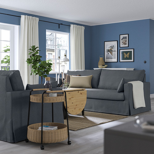 Acogedora sala de estar con un gran sofá HYLTARP gris oscuro, mesa de centro de madera y mesa auxiliar de mimbre contra ventanas azules con cortinas.
