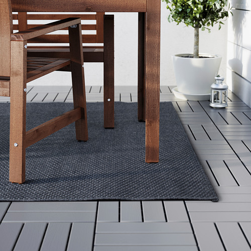 Alfombra de exterior oscura debajo de muebles de patio de madera. Tarima gris y jardinera blanca a la vista.