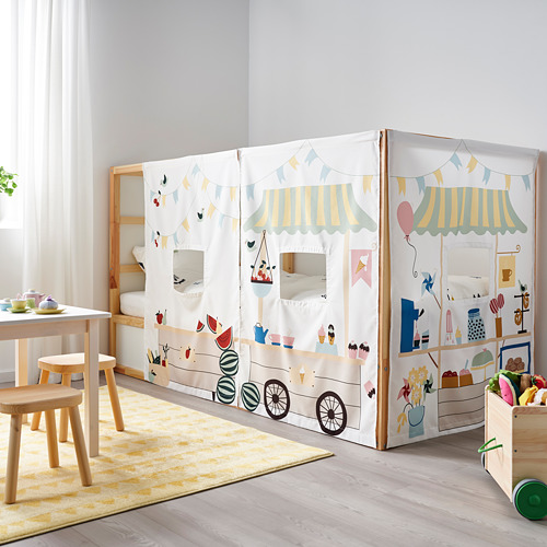 Cama blanca para niños con carpa de colores, mesa de madera, sillas y papelera de juguetes en una habitación luminosa.