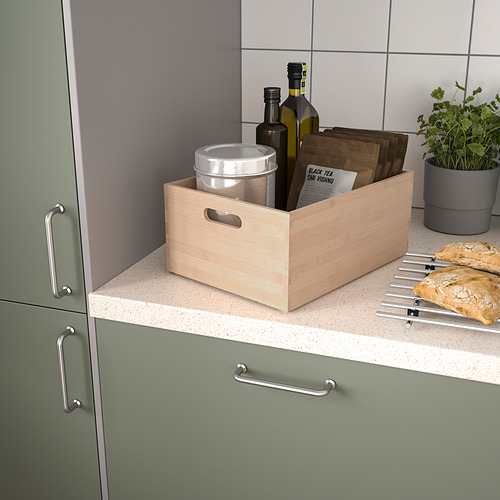 Caja de madera clara en el mostrador entre armarios verdes que contiene botellas, lata, tablas de madera, hierbas.