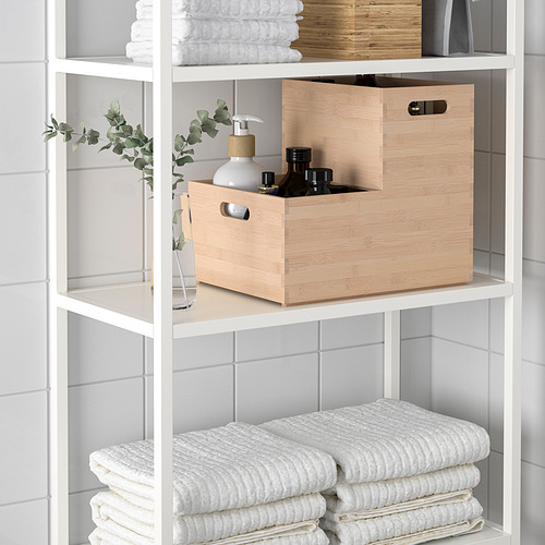 Estantes blancos con toallas dobladas, una caja de almacenamiento de madera clara con botellas negras en su interior y una pequeña planta en maceta.