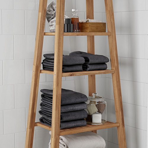 Estantería de baño de madera con tres niveles, con toallas oscuras dobladas, artículos de tocador y un frasco de vidrio, ideal para organizar los elementos esenciales del baño.