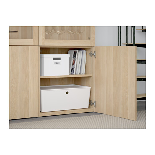 Armario de madera abierto que muestra el almacenamiento organizado. El estante inferior tiene un gran contenedor blanco; El estante superior contiene una caja blanca y revistas. Puertas de vidrio arriba.