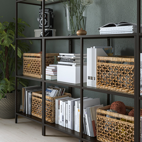 Estantería de metal negro con estantes de vidrio, que exhibe libros, cestas y artículos decorativos. Pies ajustables para mayor estabilidad en suelos irregulares.