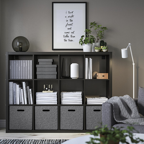 Estantería kallax negra con cajas de almacenamiento de fieltro BLÄDDRA, libros, plantas, arte enmarcado, lámpara, sofá gris y alfombra gris, organizada y ordenada.
