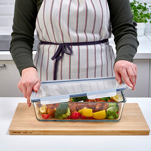 Persona que coloca la tapa en un recipiente de vidrio para alimentos lleno de pollo cocido, brócoli y rodajas de cítricos, que descansa sobre una tabla de cortar de madera de ikea.
