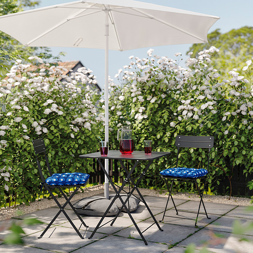 Pequeño patio con mesa plegable y sillas acolchadas azules, a la sombra de una sombrilla blanca, frente a una pared de flores blancas en flor.