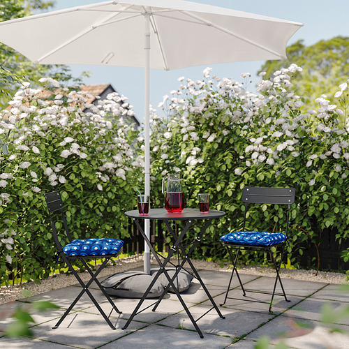 Pequeño patio exterior con mesa y sillas plegables, bajo una sombrilla blanca, rodeado de arbustos de flores blancas.