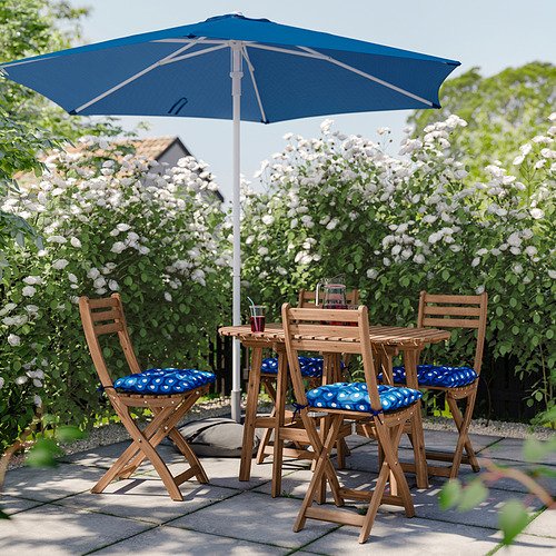Entorno exterior con mesa y sillas de madera de color marrón claro, cojines azules y sombrilla azul a juego, rodeado de exuberantes flores blancas.