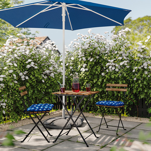 Patio al aire libre con mesa, sillas y cojines azules bajo una sombrilla azul, rodeado de flores blancas.