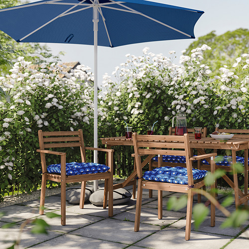 Patio al aire libre con mesa y sillas de madera de acacia, cojines azules y sombrilla a juego rodeada de flores blancas.