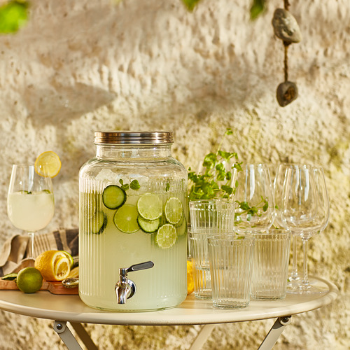 Dispensador de bebidas de vidrio transparente con grifo de plata, rodajas de lima, vasos, en soporte de madera contra pared de piedra.