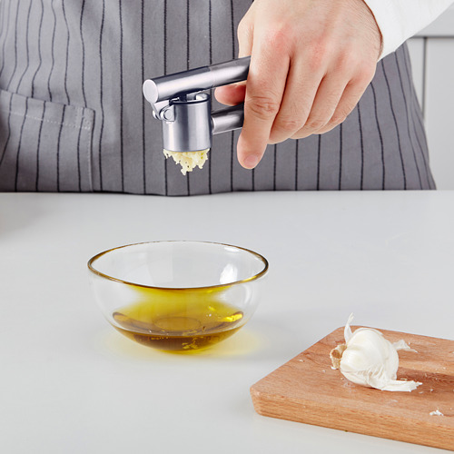Persona prensando ajo con una prensa de ajo KONCIS, picando clavos en un recipiente de vidrio con aceite sobre una encimera blanca.
