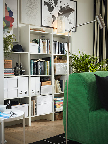 Estantería blanca con libros y decoración, sillón verde, planta.