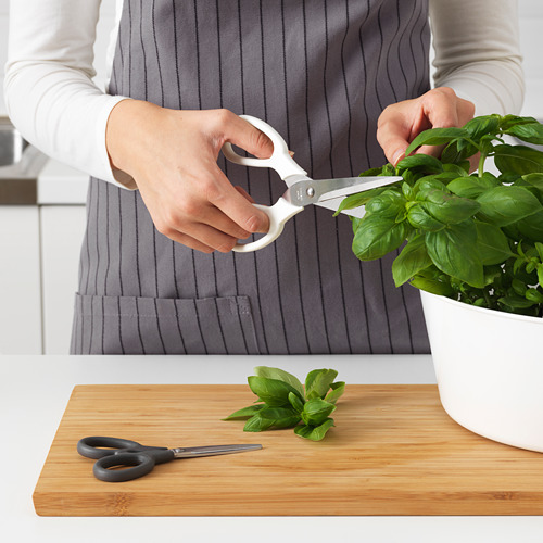Una persona sostiene unas tijeras blancas cortando albahaca fresca sobre una tabla de cortar de madera con un par de tijeras adicionales y hojas de albahaca cortadas en ella.