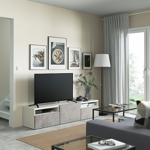 Sala de estar moderna con banco de televisión BESTÅ, TV de pantalla plana, arte, cortinas grises, sofá acogedor y mesa de centro de vidrio cerca de una gran ventana.