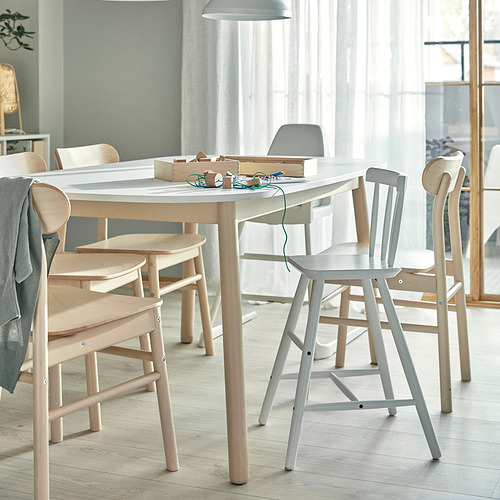 Mesa de comedor moderna con sillas de madera clara. Manualidades, herramientas y papel sobre la mesa. Gran ventanal con cortinas transparentes. Decoración minimalista, habitación espaciosa.