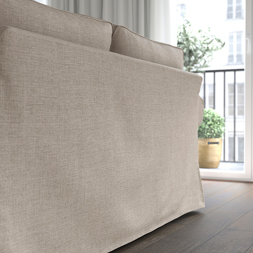 Sofá tapizado en color beige cerca de ventana con planta. Cojines suaves y reversibles para sentarse cómodamente.