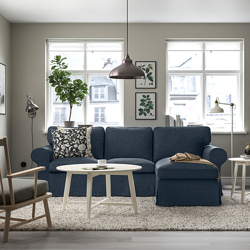 Acogedora sala de estar con sofá azul, otomana y mesa de café. Grandes ventanas, alfombra gris, lámpara, silla y lámpara cálida.