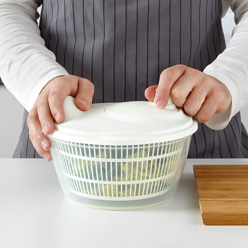 La persona sostiene una centrifugadora de ensaladas blanca UPPFYLLD, enjuagando la ensalada en el fregadero de la cocina. La centrifugadora seca la ensalada, con agua acumulada en el recipiente.