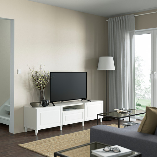 Sala de estar moderna con mueble de TV blanco BESTÅ. Tv negra, jarrón con ramas, cortinas grises. Suelo de madera oscura, alfombra clara.