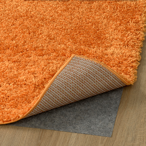 Alfombra peluda de color naranja brillante con almohadilla antideslizante gris texturizada debajo, colocada sobre piso de madera.