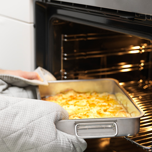 Una persona con guantes de horno saca una fuente de asar llena de comida de un horno. La lata tiene asas y es apta tanto para uso en horno como en parrilla.