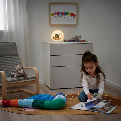 Una niña lee un libro sobre una alfombra de colores en una habitación acogedora. La alfombra tiene un diseño de erizo. Un cofre cercano contiene un animal de peluche y una lámpara encendida.