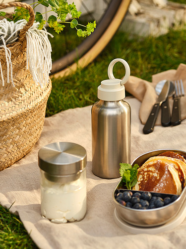 Una botella de agua de acero inoxidable con tapa y asa beige, junto a un frasco de yogur y un tazón de bayas, colocada sobre una manta de picnic.