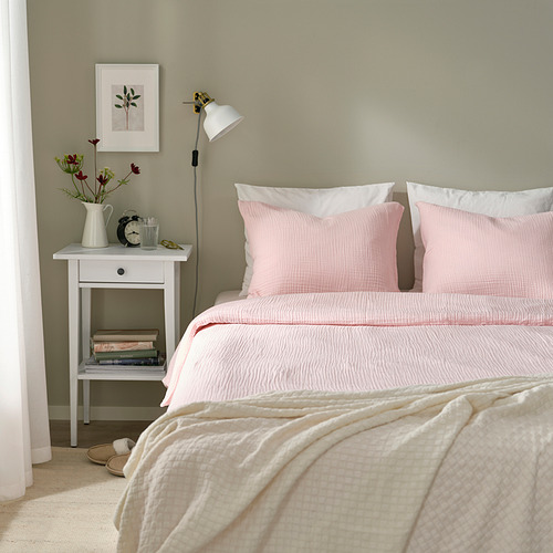 Cómoda cama con almohadas rosas y edredón SCHERSMIN, mesita de noche pequeña con lámpara y flores.