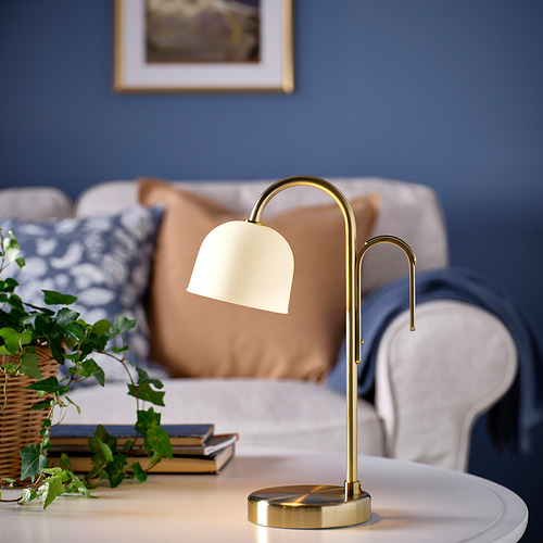 Lámpara TVÅMASTAD sobre mesa con sofá y planta. Lámpara dorada con pantalla de cristal esmerilado y mango portátil.