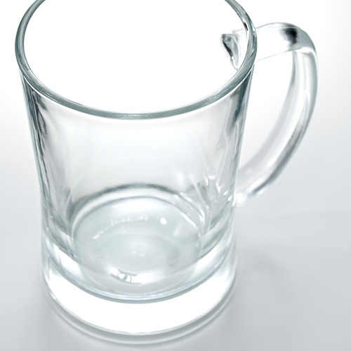 Una jarra de vidrio transparente y resistente con un asa ancha se encuentra vacía sobre una superficie reflectante, ideal para servir cerveza con la cabeza llena.